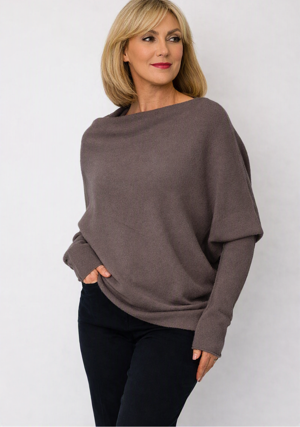 A woman wearing a loose, oversized long-sleeve knit sweater in taupe brown with black pants, standing against a light gray studio background. 